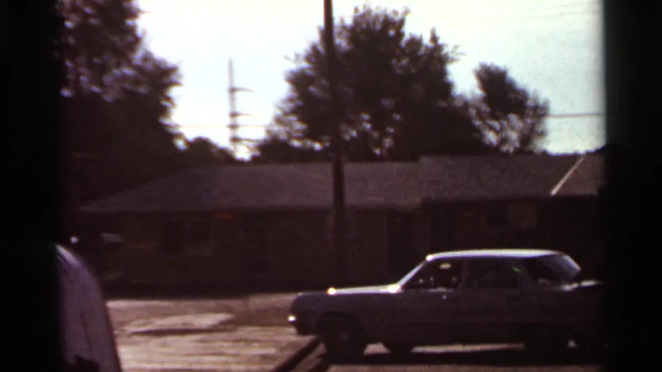 1966 St. Louis roadside motel scene with a vintage car in front, low building, trees, pavement, and utility lines in grainy 8mm color.