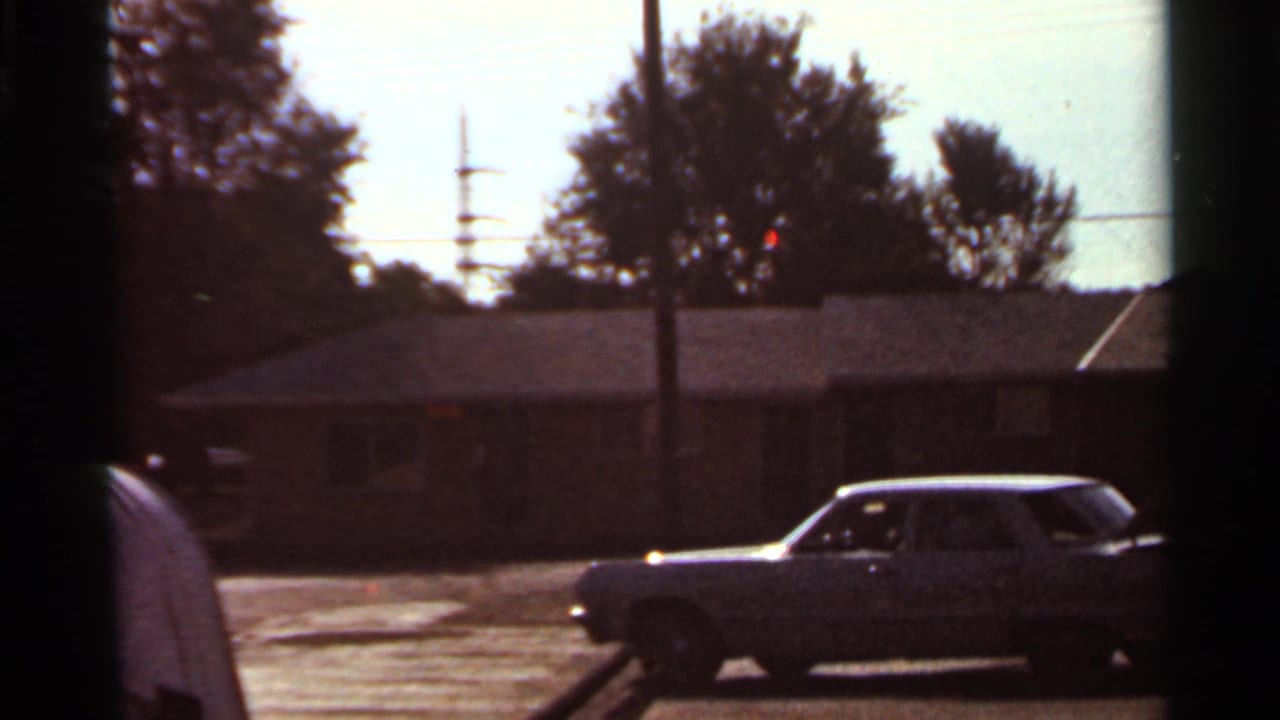 1966 Saint Louis home-movie frame showing a classic sedan in a residential parking lot with brick buildings and a person partly at left.