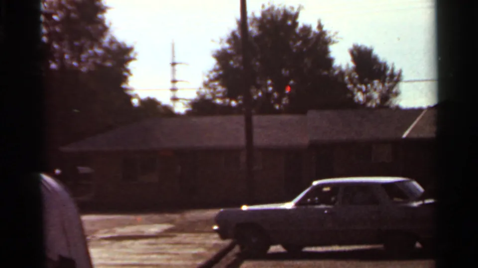 1966 Saint Louis home-movie frame showing a classic sedan in a residential parking lot with brick buildings and a person partly at left.