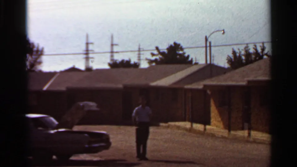 A man stands farther from the open-hood sedan in a 1966 Saint Louis residential parking lot, with brick buildings and power lines visible.