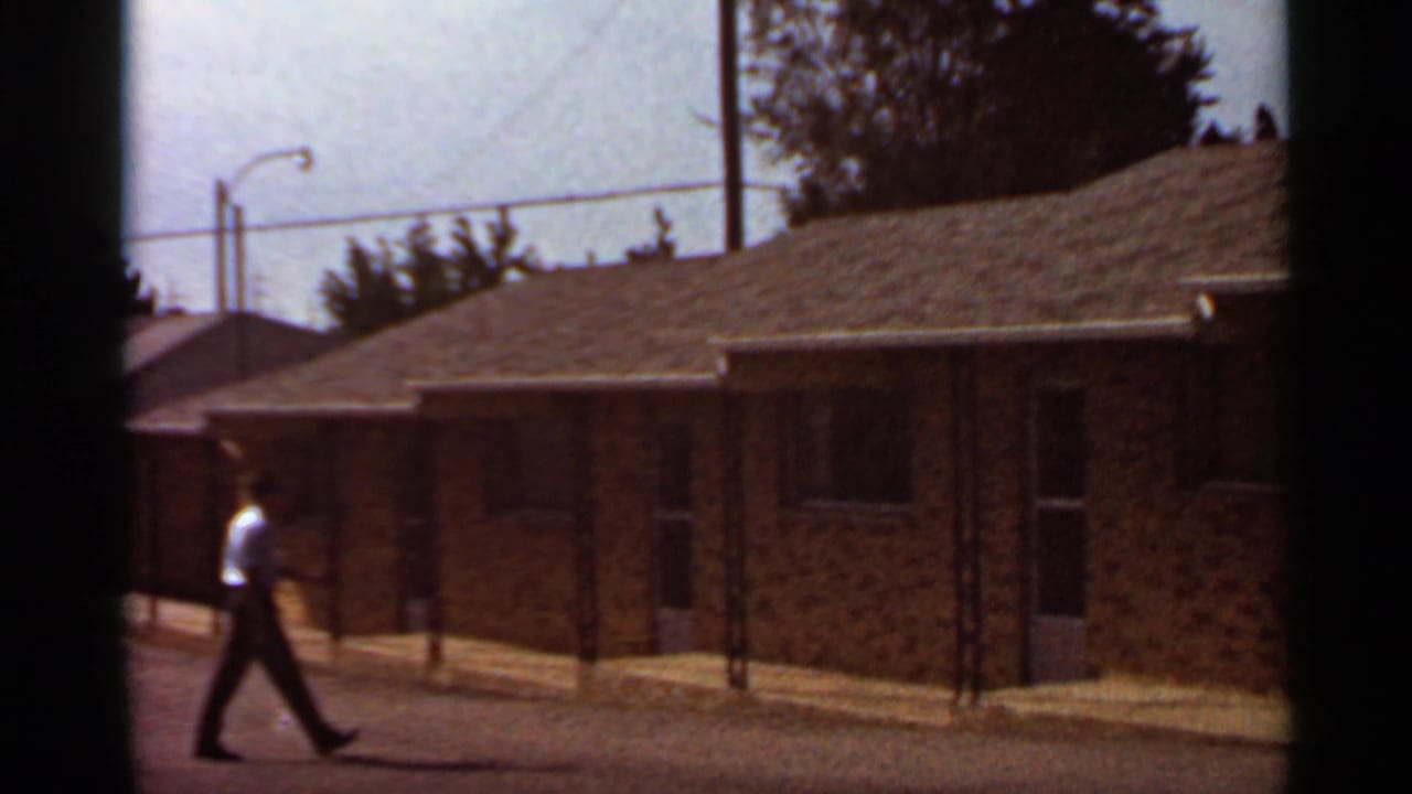 Adult in a light shirt walks past brick motel rooms under a bright sky.