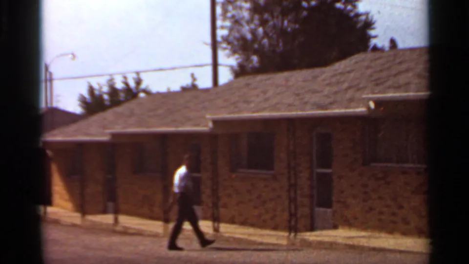 The adult continues along the Saint Louis motel exterior, passing brick walls, doors, posts, and a sunlit sidewalk.