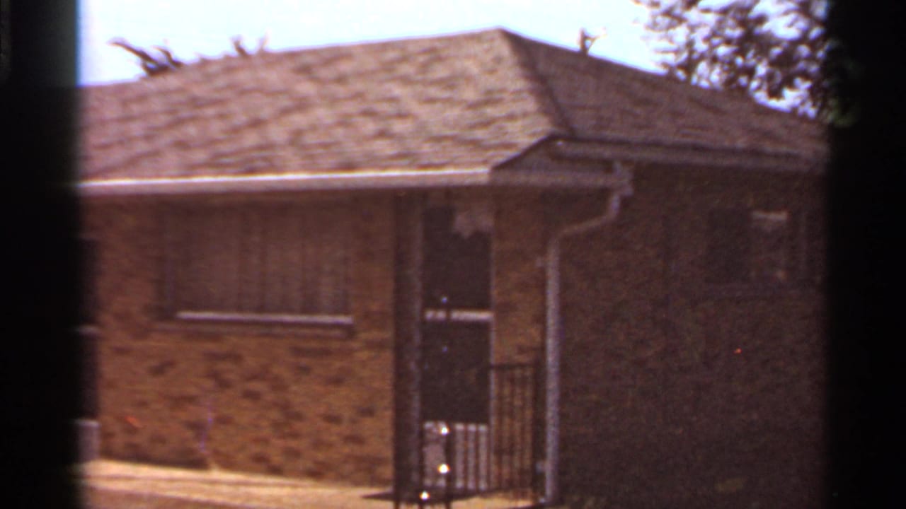 Brick apartment building with a doorway, small windows, roofline, and shaded trees behind it.