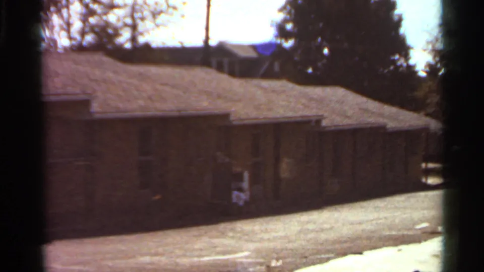 1966 Saint Louis home-movie frame showing a long low brick apartment row beside a bright driveway with trees behind it.