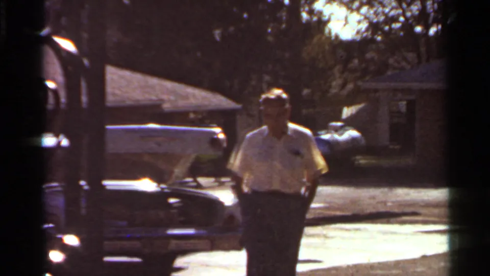 1966 Saint Louis home-movie frame showing an adult man walking past a parked car near modest residential houses.