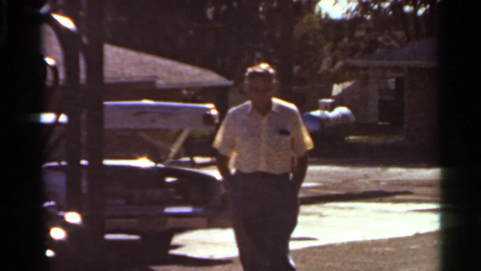 Adult man in a light shirt walks near a parked car on a Saint Louis residential street in 1966 home-movie footage.