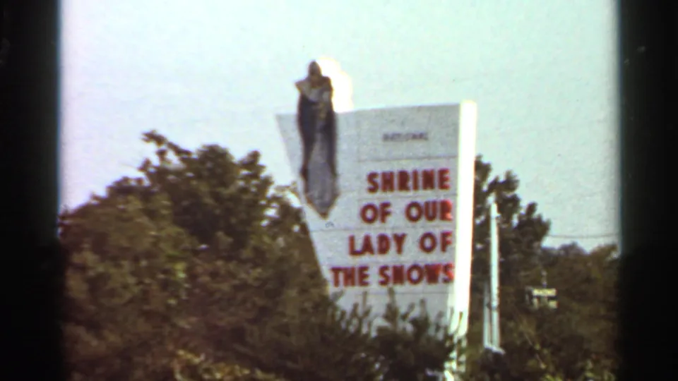1966 Saint Louis-area frame showing the Shrine of Our Lady of the Snows roadside sign with a robed statue form above trees.