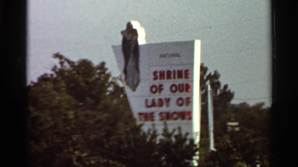 1966 Saint Louis-area frame with a cropped view of the Shrine of Our Lady of the Snows sign partly obscured by trees.