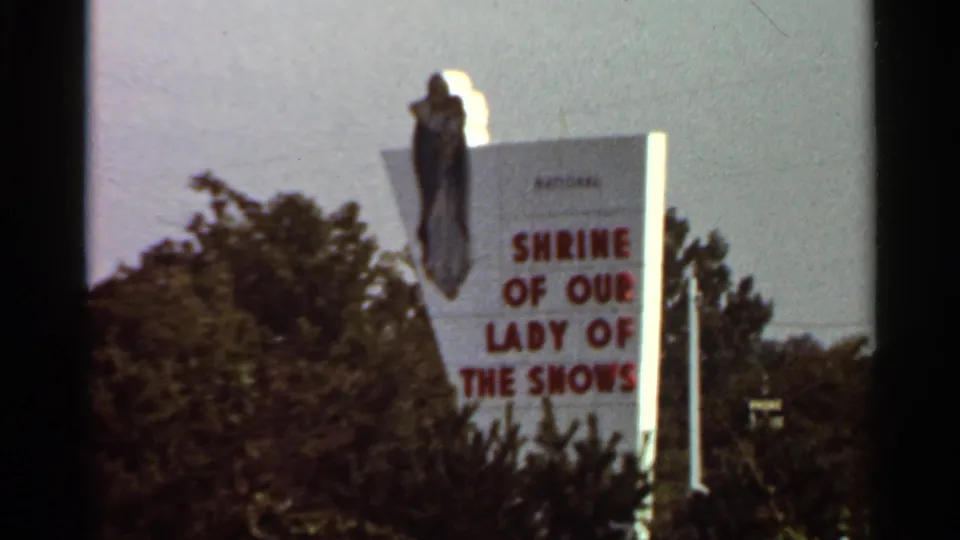 1966 Saint Louis-area frame showing a steadier view of the Shrine of Our Lady of the Snows roadside sign above trees.