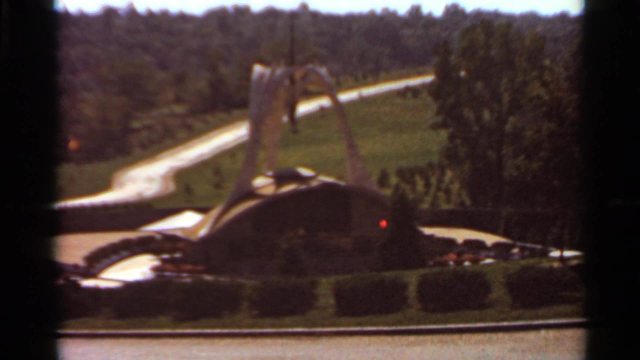 1966 Saint Louis 8mm frame of the curved decorative fountain and central spire against rolling green grounds and trees.