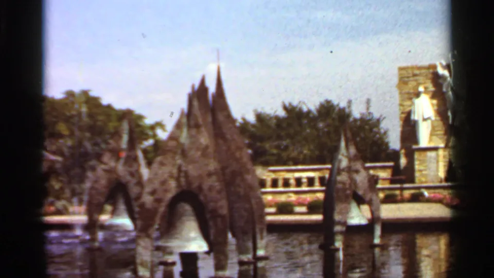 1966 Saint Louis home-movie frame showing sculptural fountain spires, water spray, a reflecting pool, trees, and a white statue on a stone pedestal.