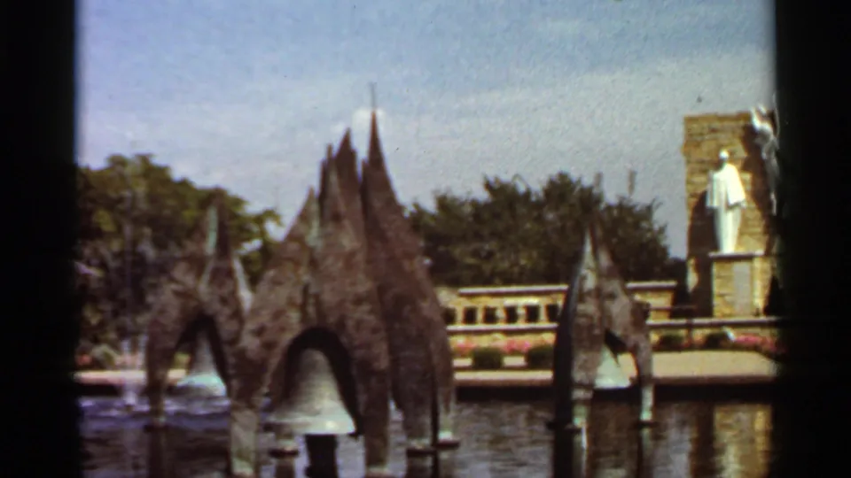 1966 Saint Louis frame with dark fountain structures in the water, pale spray or bell-shaped water, a stone backdrop, trees, and a distant white statue.