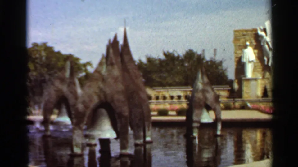 1966 Saint Louis frame showing several dark fountain forms across reflective water, with trees, low railings, a stone monument, and a white standing statue beyond.