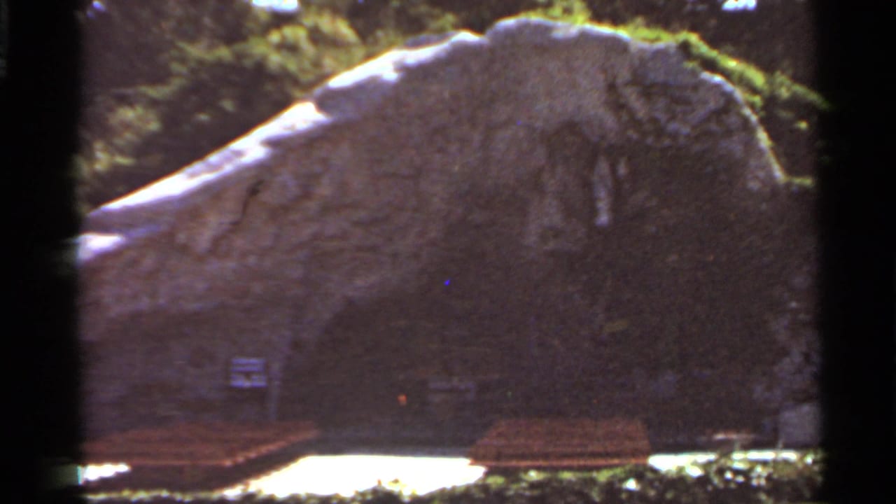 1966 Saint Louis 8mm frame showing a wooden bench, bright grass, shrubs, and a large rock face with a dark opening.