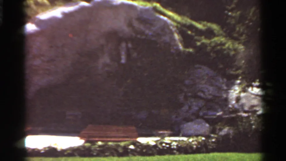 1966 Saint Louis 8mm still with a bench at the base of a dark rock face, bordered by lawn and foliage.
