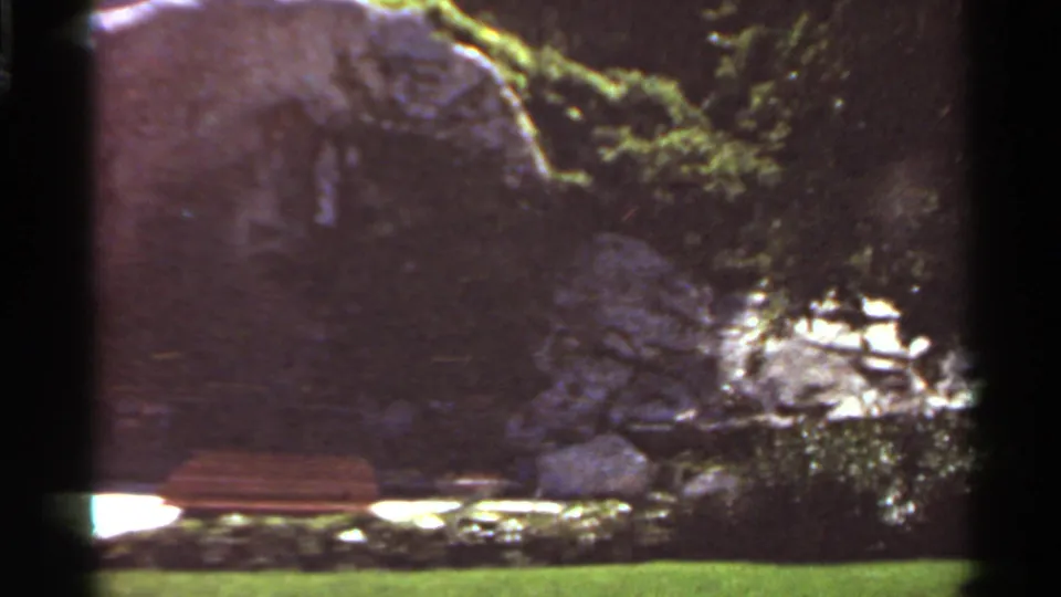 1966 Saint Louis frame showing a quiet bench and green lawn near a shadowed rock formation and surrounding shrubs.
