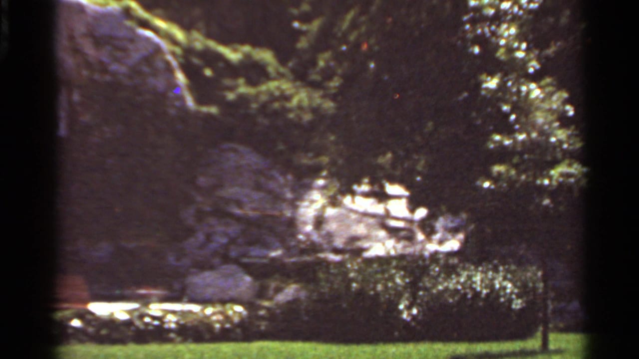 1966 Saint Louis footage frame of a natural rock landmark surrounded by greenery, benches, lawn, and small signs.