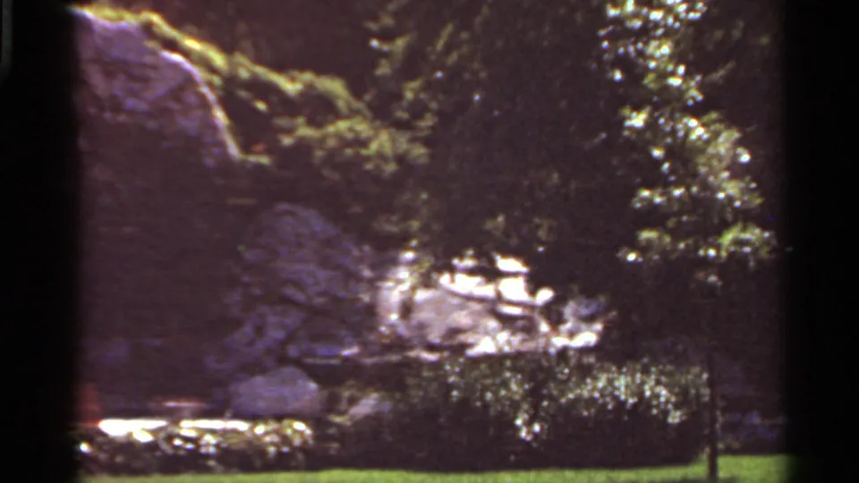1966 Saint Louis 8mm frame showing a boulder formation partly shaded by trees with grass, plantings, and a bench nearby.