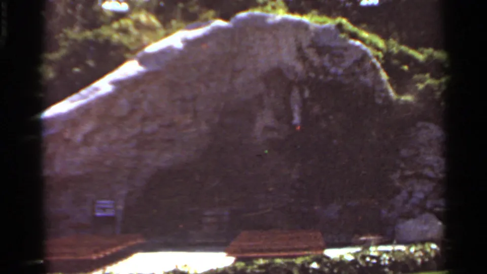 1966 Saint Louis home-movie frame showing a stone grotto, benches, shrubs, and bright garden paving around a religious shrine.