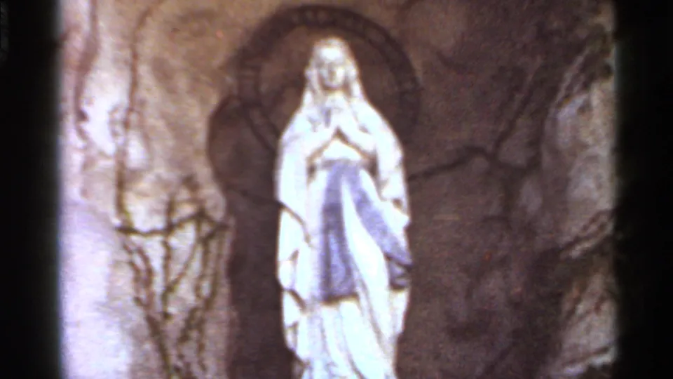 1966 Saint Louis 8mm frame of a white Marian statue inside an arched grotto niche, with bare vines and stonework around it.