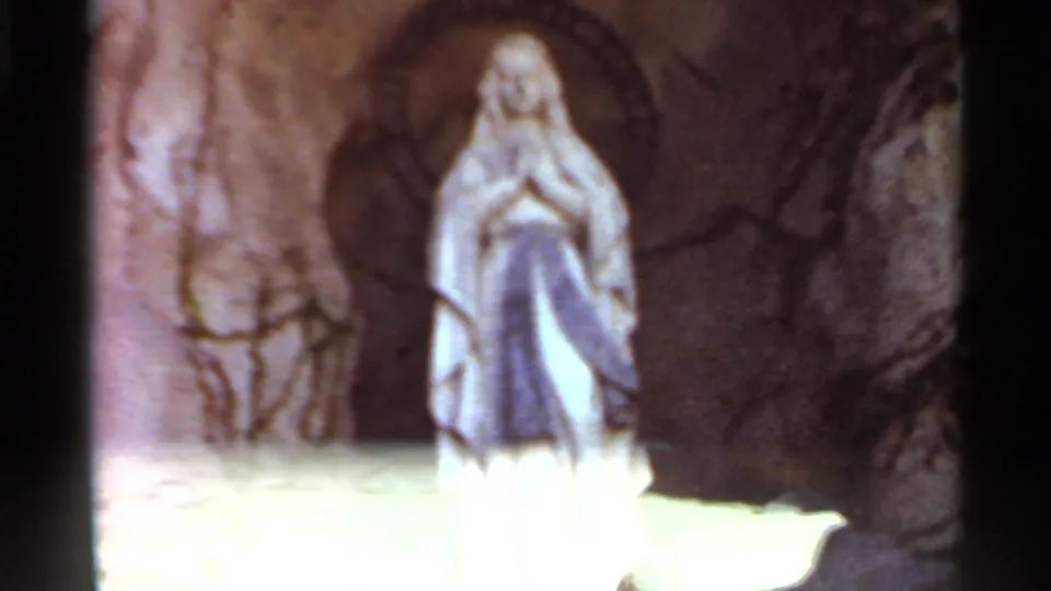 1966 Saint Louis frame showing the Marian statue against a dark arched grotto, with bright patio paving below and vines at the side.