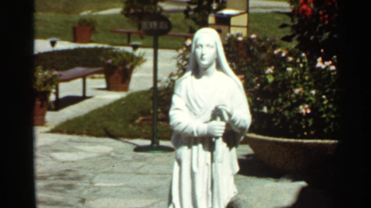 1966 8mm frame of a white robed female statue in a landscaped garden, with stone paving, lawn, and potted plants around it.