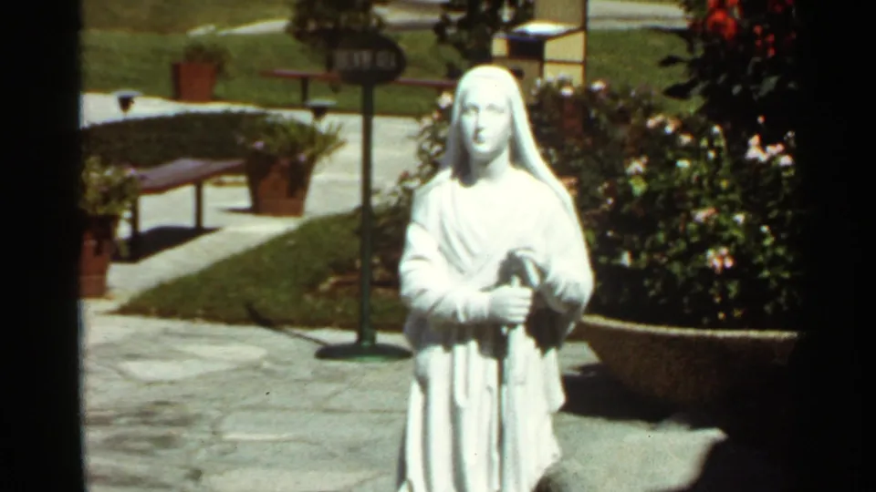 1966 8mm frame of a white robed female statue in a landscaped garden, with stone paving, lawn, and potted plants around it.