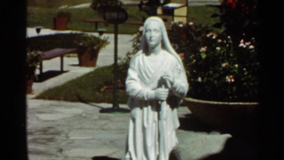 The 1966 Missouri home-movie frame holds on a white robed female statue beside flowers, paving, and a low garden planter.
