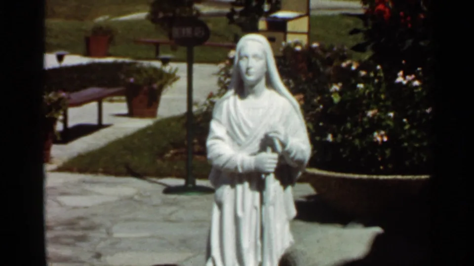A 1966 8mm view of the same white garden statue, seen with stone paving, lawn, flowers, and visitor-area plantings.