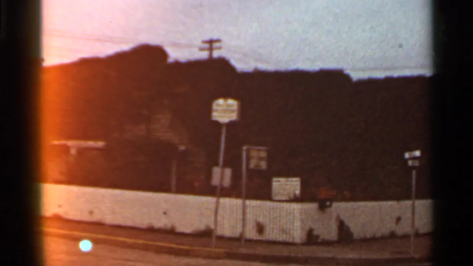 1966 St. Louis home-movie view of a white fence, multiple roadside signs, utility poles, and a dark wooded or hillside backdrop.