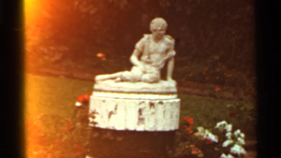 1966 St. Louis frame showing a weathered seated figure statue on a pedestal in a small garden with flowers and grass.