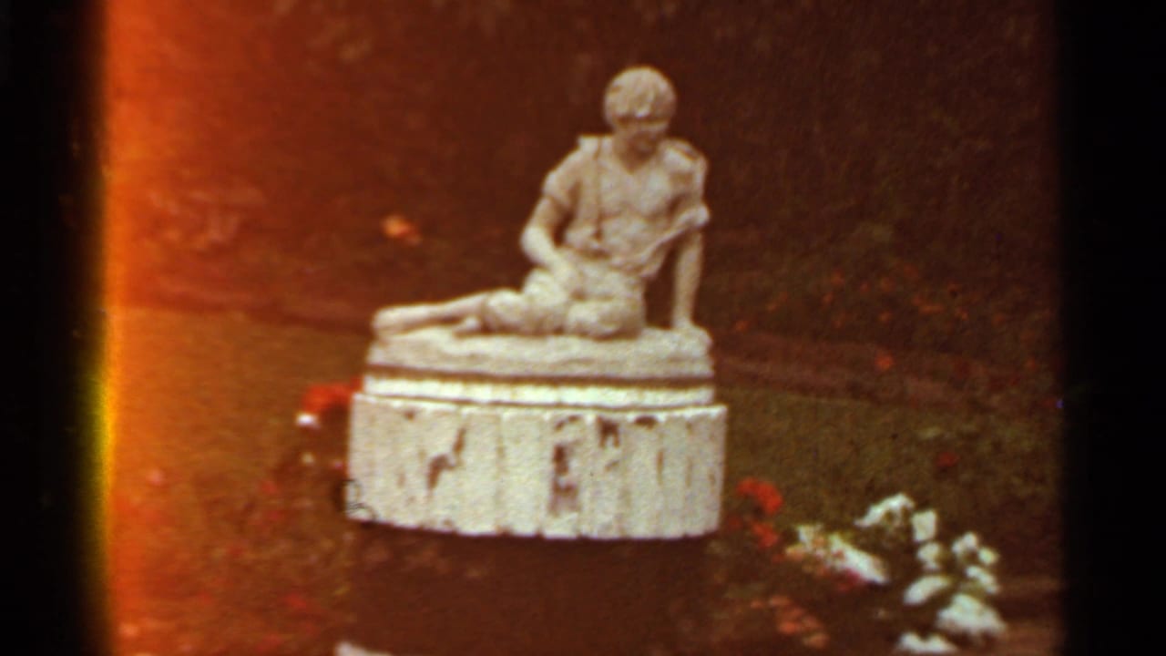Seated stone statue on a pedestal beside flowers in a landscaped outdoor setting.