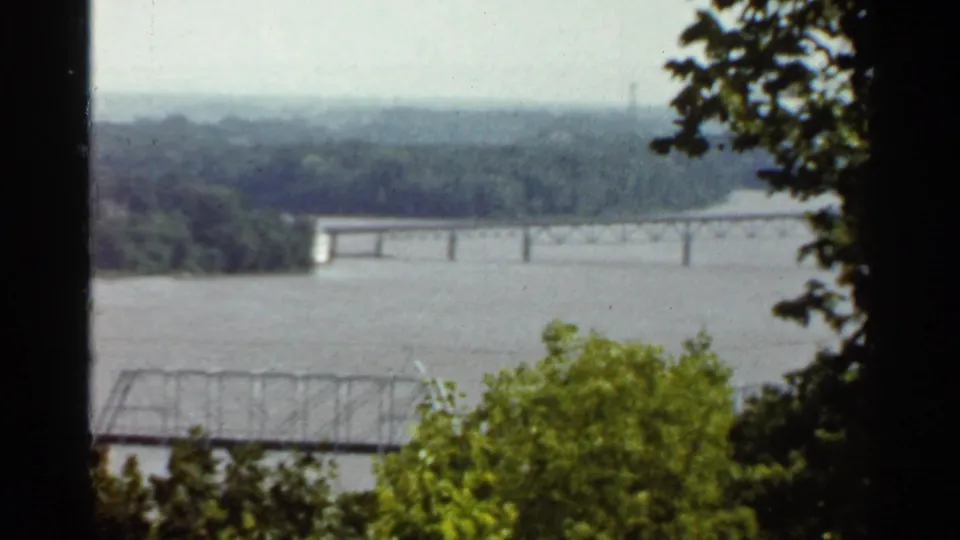 1966 Saint Louis home-movie frame of bridges over a broad river, partially obscured by foreground foliage.