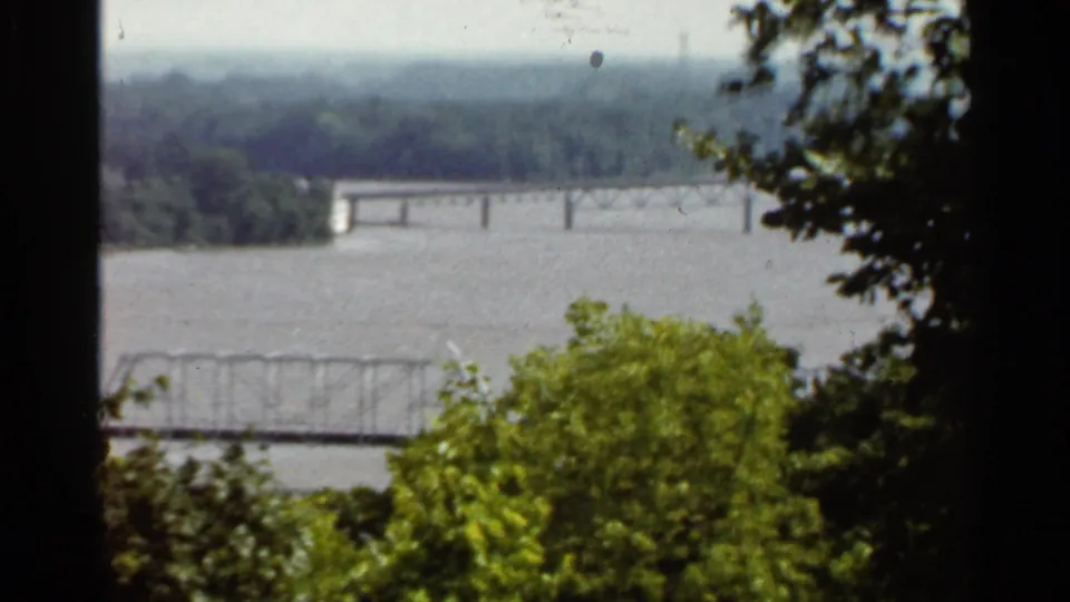 1966 Saint Louis river overlook with distant bridge spans, green foreground trees, and soft 8mm grain.