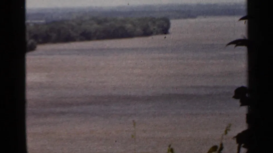 Saint Louis-area 1966 home-movie frame showing a broad river from above, with distant trees and dark foreground plants at the edge.