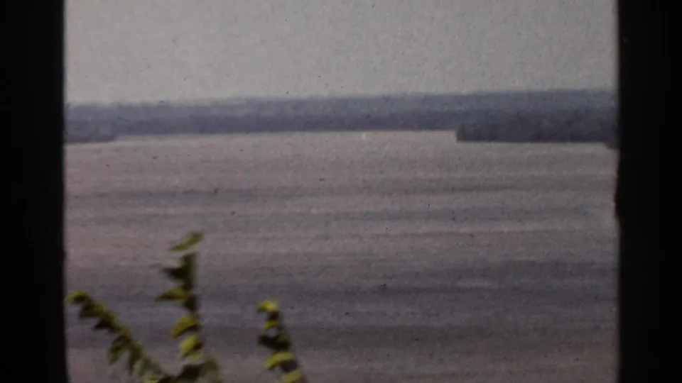 Final 1966 8mm frame of the Saint Louis river overlook, with a broad waterway, distant wooded shore, and blurred foreground greenery.