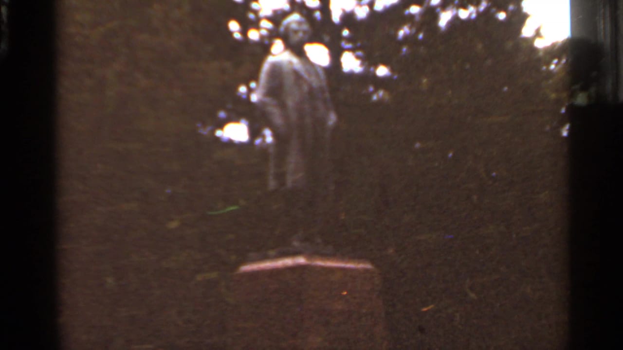 1966 Saint Louis frame showing a Mark Twain statue on a stone pedestal amid trees, shot in soft faded 8mm color.