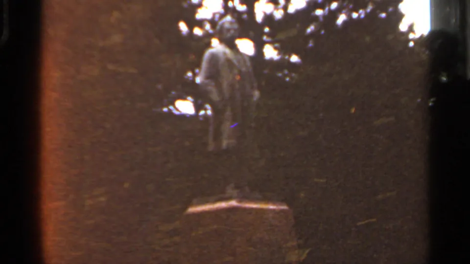 1966 Saint Louis 8mm frame showing a distant outdoor statue on a pedestal, seen through shadowed trees.