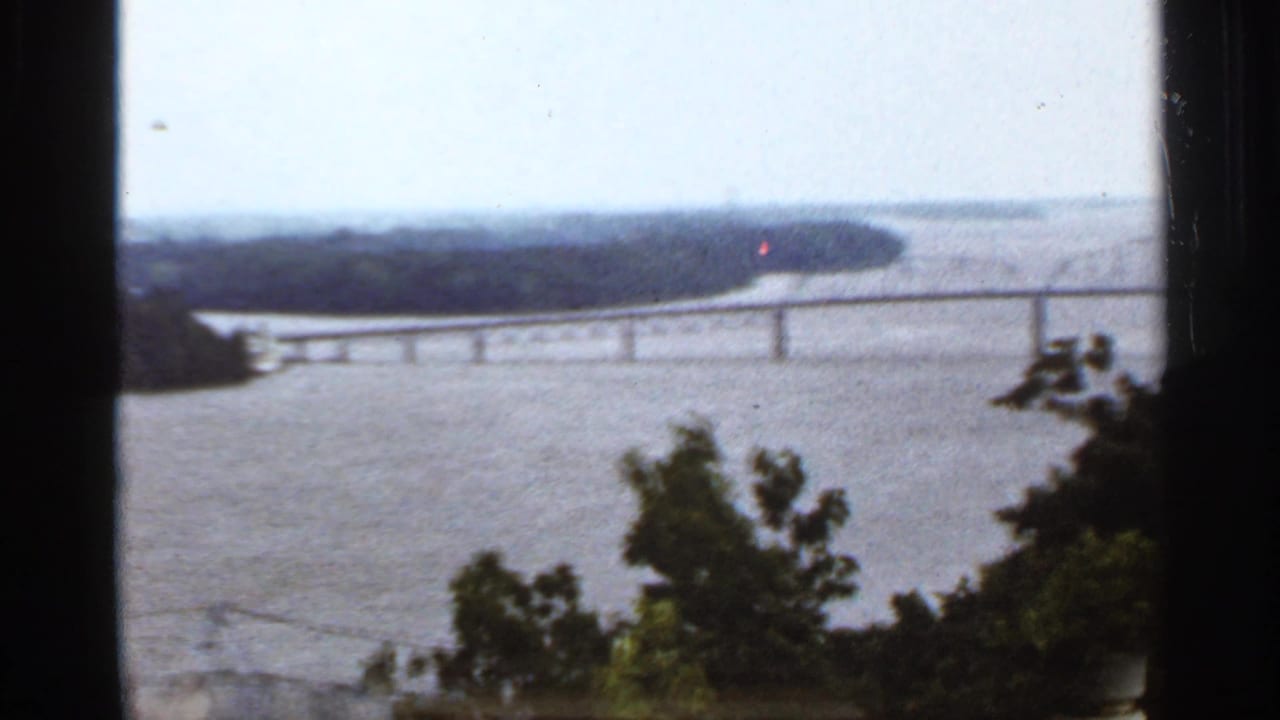 1960s 8mm home-movie view of a broad river and long bridge framed by dark foreground trees.