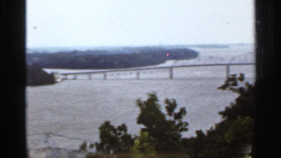 1960s 8mm home-movie view of a broad river and long bridge framed by dark foreground trees.