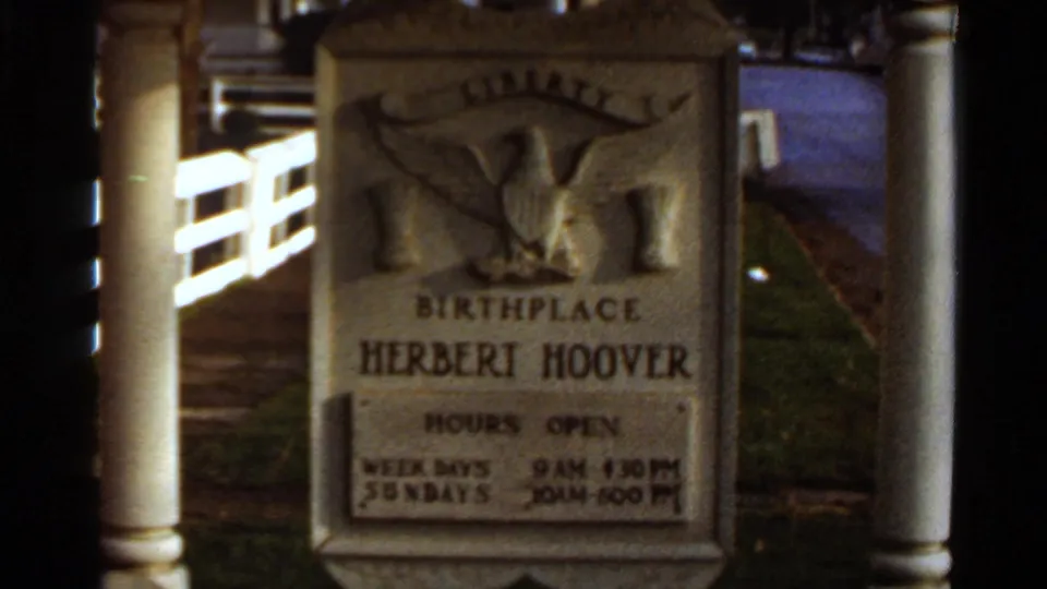 Stone Herbert Hoover birthplace sign with eagle relief, posted hours, white fence, and walkway in 8mm footage.