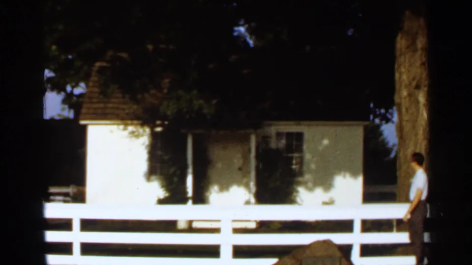 Small white historic house behind a white rail fence, with trees overhead and an adult visitor walking at right.