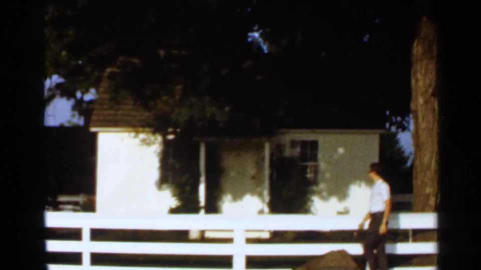 1966 8mm frame showing an adult man near a small white house and white fence, with trees casting shade around the scene.