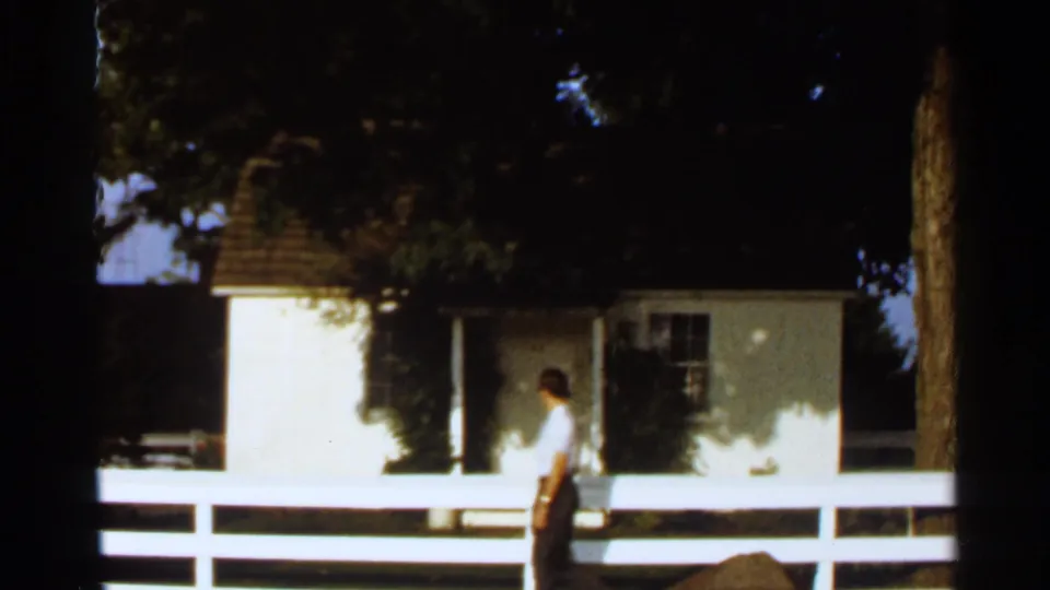 1966 8mm frame of an adult man beside a white fence, with a small white house, porch area, and trees in the background.