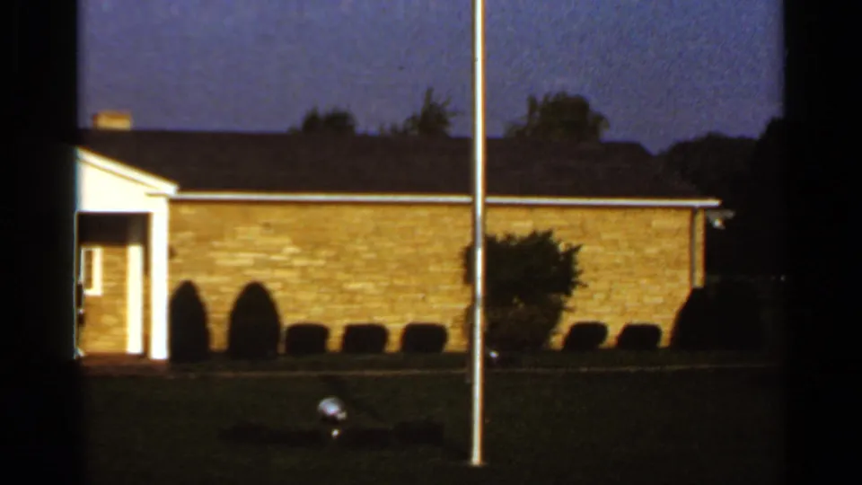 Visitor-shot 1966 exterior view of a low brick institutional building at dusk, with lawn, shrubs and a flagpole in the foreground.