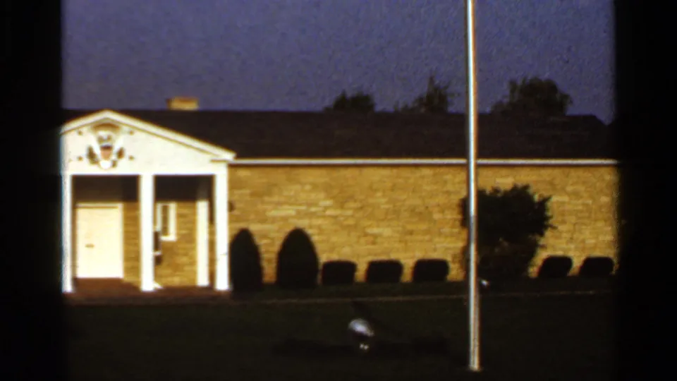 1966 8mm exterior frame of the Herbert Hoover Presidential Library building, showing white columns, a lit entrance area, shrubs and a flagpole.