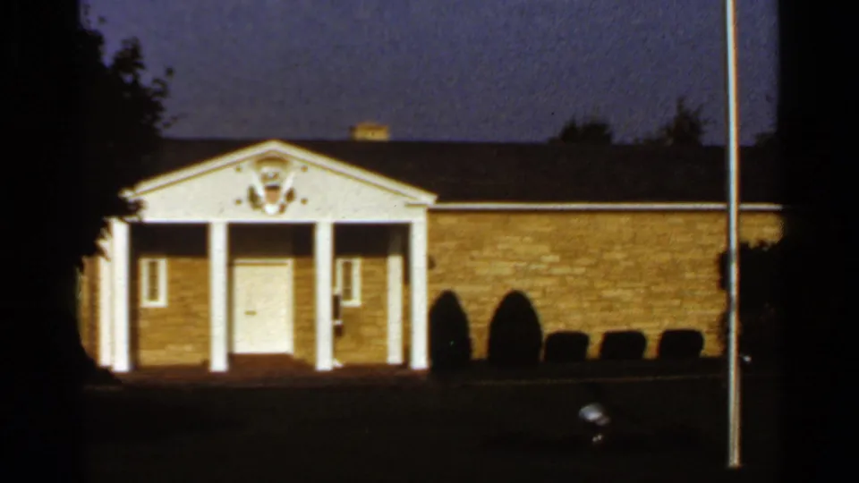 1966 home-movie frame centered on the library entrance, with a white-columned portico, brick wall, flagpole and deep dusk shadows.