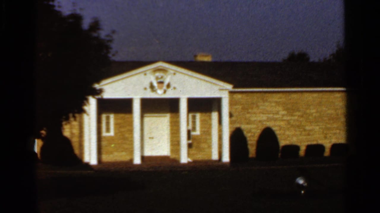 1966 St. Louis home-movie frame showing a brick building with white columns, a central door, and a crest above the entrance at dusk.