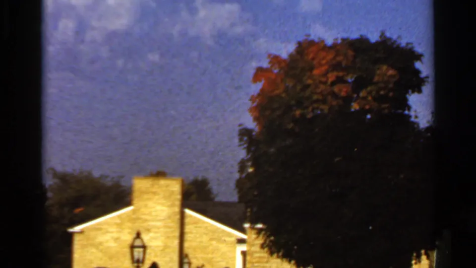 1966 St. Louis-area home-movie frame with a brick house, chimney, lawn lamp, and large wind-darkened tree canopy.