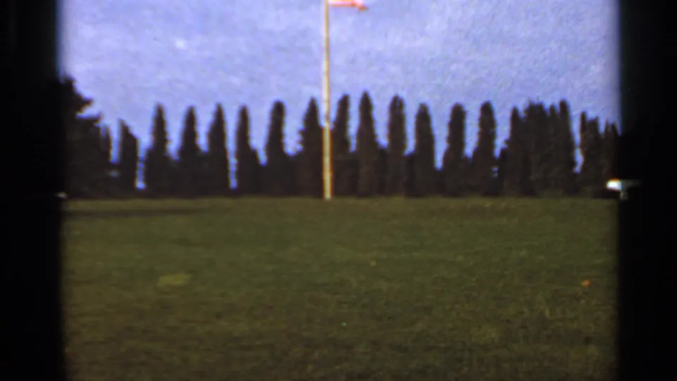 1966 St. Louis-area 8mm frame showing a grassy field, flagpole, cloudy sky, and a border of tall evergreens.